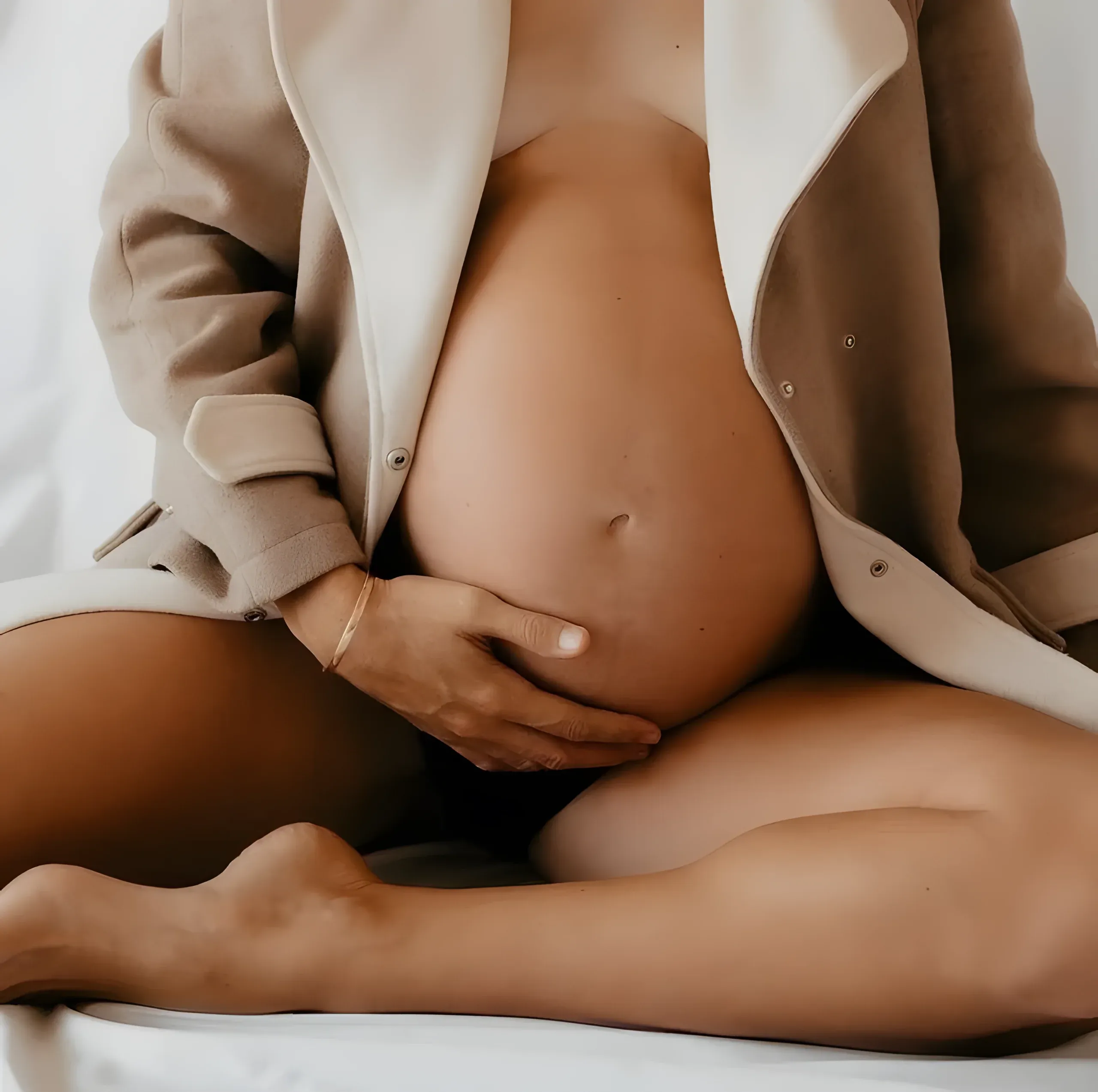 Femme enceinte assise, manteau beige ouvert, une main posée sur son ventre