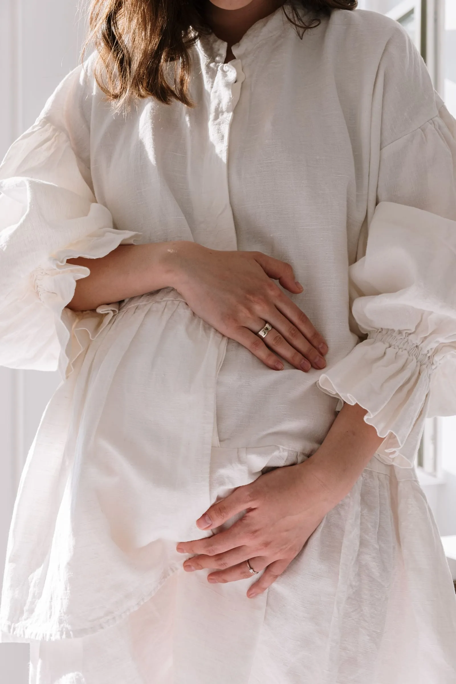 Femme enceinte en tenue de lin blanc cassé, les mains croisées sur son ventre