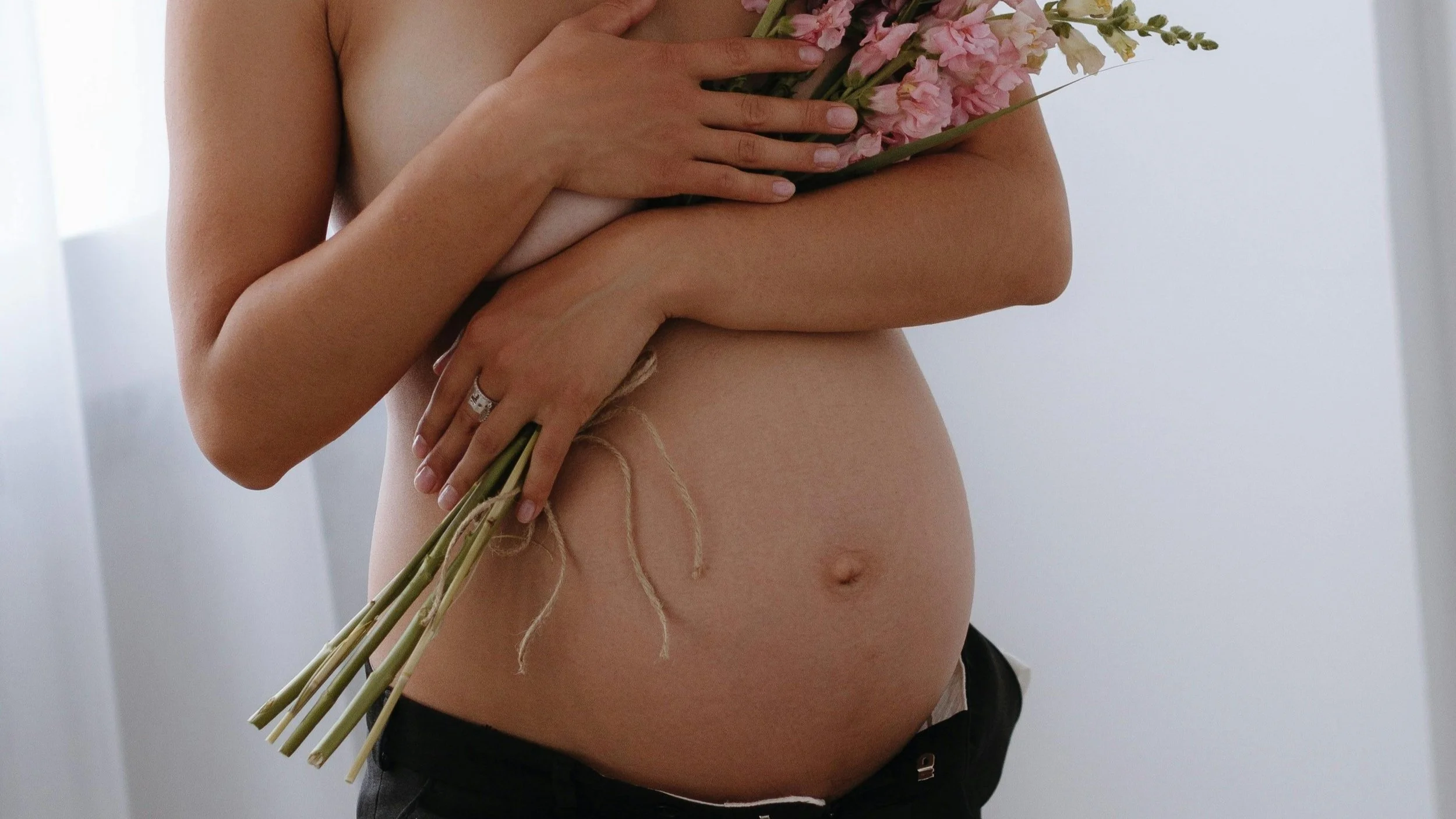 Femme enceinte tenant un bouquet de fleurs roses contre son ventre nu, dans une pièce baignée de lumière